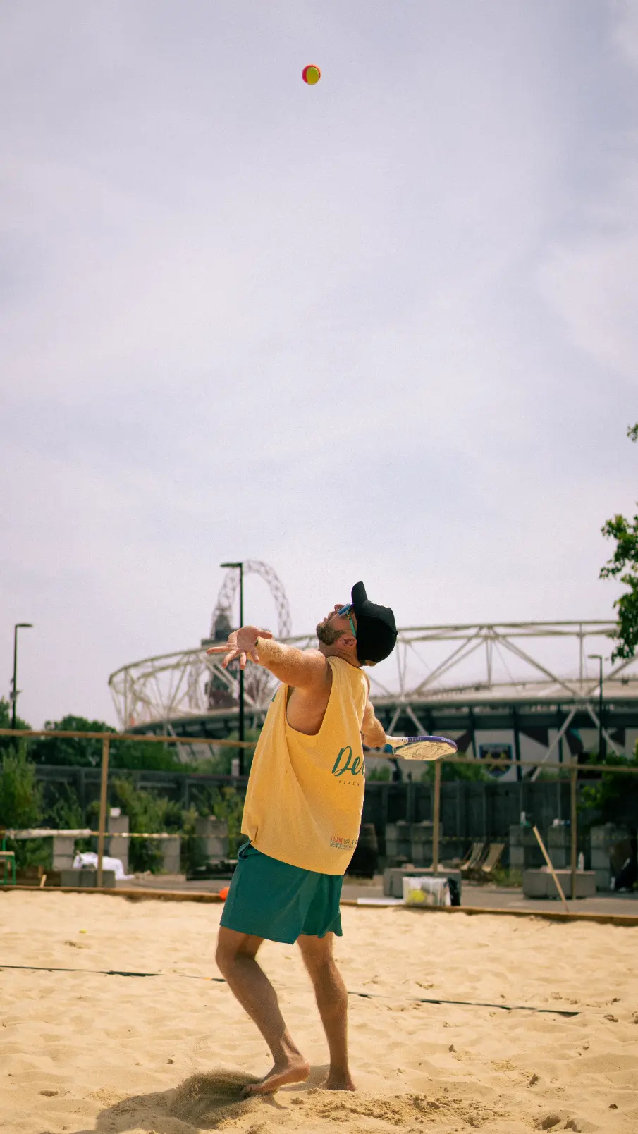 A person playing beach tennis prepares to hit a ball high in the air. They are wearing a yellow tank top, green shorts, and a black baseball cap. London Stadium in the Olympic Park is visible in the background.