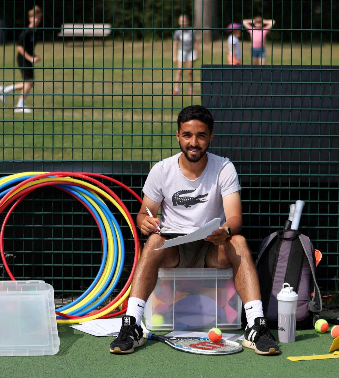 A person is sitting on a plastic container in a tennis court, holding a clipboard and looking at the camera. A tennis racket and balls, hoops, and other sports equipment surround them