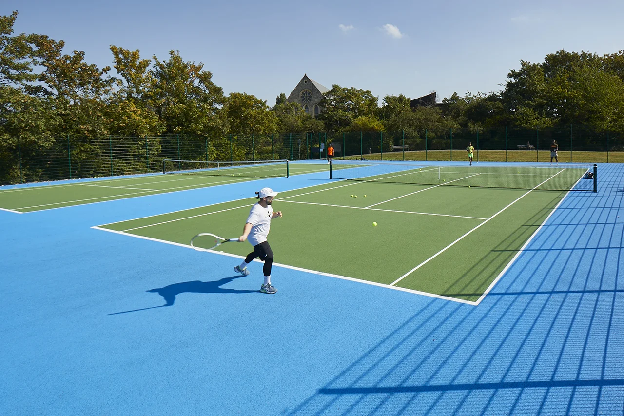 A person in a white t-shirt and baseball cap swings a racket at a yellow tennis ball on a vibrant blue and green outdoor court on a sunny day